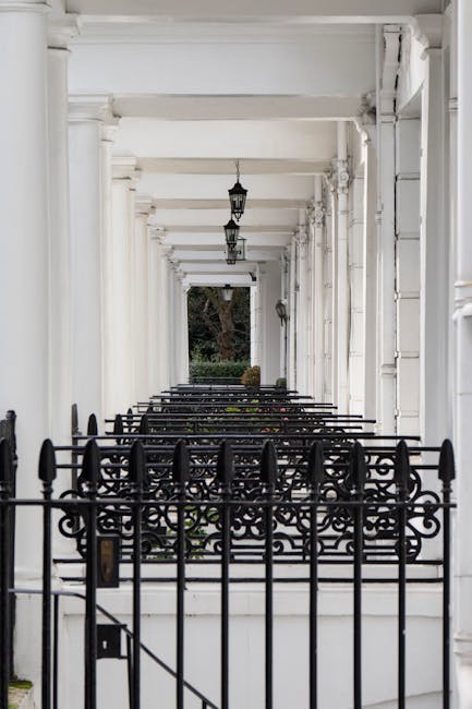 A long, covered white porch with decorative white columns and a series of hanging black lantern-style lights. The porch features a black wrought iron railing with intricate scrollwork design in the foreground, separating the walkway from the garden area. Beyond the railing, green trees and shrubbery are visible at the end of the porch. The surface is clean, with a smooth white flooring that appears well-maintained, reflecting light in some areas, indicating recent surface cleaning or polishing. The overall scene exudes a neat, pristine appearance typical of property maintenance in South Kensington. The setting represents a stylish, well-maintained residential exterior, matching the theme of high-quality domestic cleaning services offered by Kensington Carpet Cleaning.