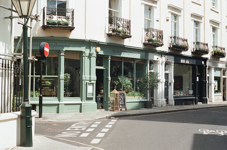 Exterior view of a light green-painted shopfront with large glass windows displaying plants within, situated on a street corner in South Kensington. The building has black wrought iron balconies on the upper floors, and a sign indicating 'Kensington Flower' on the side. A blackboard sign is placed outside the entrance, and the sidewalk in front is clean with a crosswalk and bicycle lane markings visible. The scene is well-lit, highlighting the tidy and inviting appearance, with Kensington Carpet Cleaning's mention indicating a focus on thorough end of tenancy surface cleaning and sanitisation services in the area.