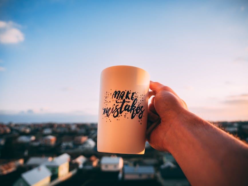 A person's hand holding a cream-colored mug with the words 'make mistakes' printed in black, casual script, against a background of an outdoor cityscape at sunset. The sky is blue with soft clouds, an