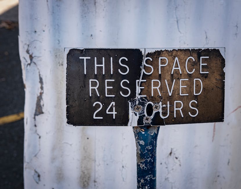 A weathered metal sign attached to a pole indicating that the space is reserved 24 hours, with peeling paint and rust visible on the sign and pole. The background features a white wall with signs of d