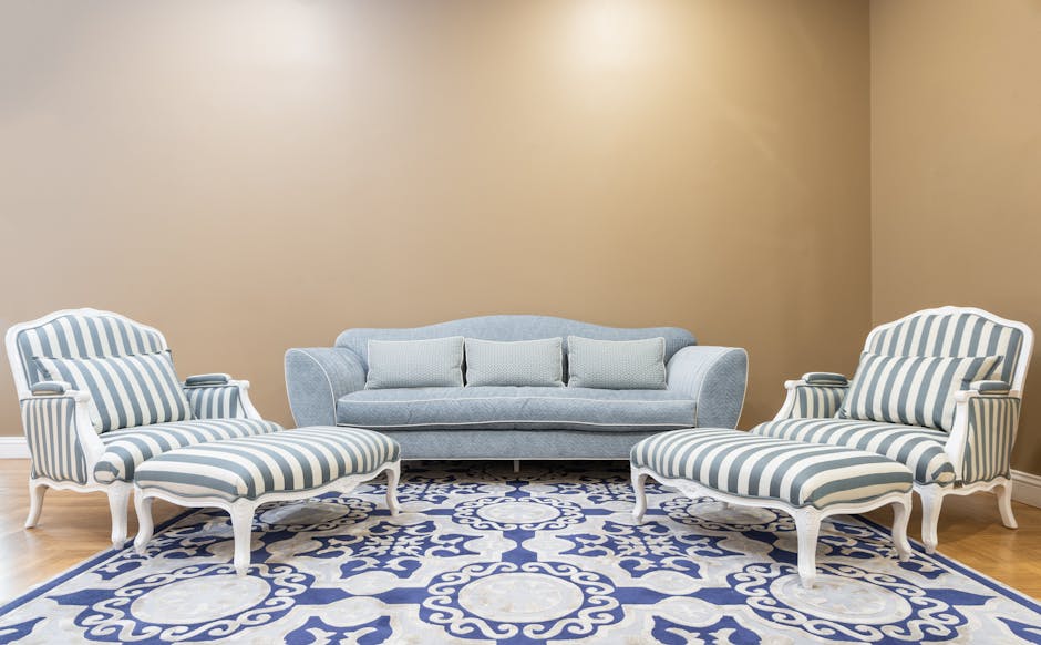 A living room with beige walls and natural light illuminating the space. In the center, there is a light blue upholstered sofa with three matching cushions, positioned against the wall. Flanking the sofa are two armchairs with white frames and blue and white striped fabric, each with matching upholstered footstools in front. The furniture is arranged on a patterned blue and white area rug that covers a light wooden floor. Visible cleaning tools or products are not present, but the room appears clean and well-maintained, reflecting surface cleaning and deep cleaning processes typical of domestic sanitisation performed by Kensington Carpet Cleaning.