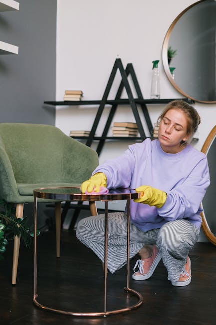 A young woman with blonde hair tied back, wearing a light purple sweatshirt, grey and white striped trousers, pink sneakers, and yellow cleaning gloves, is kneeling on the dark wooden floor of a modern living room. She is dusting or polishing a small, round glass table with a metal frame, which is in front of a green upholstered armchair. Behind her, there is a black shelf decorated with books arranged vertically and horizontally, a small potted plant, and a white spray bottle. The wall is painted light grey, and a large round mirror with a wooden frame hangs above the shelf, reflecting part of the room. The room is well-lit, emphasizing the clean and tidy appearance of the space, consistent with deep surface cleaning or home sanitisation offered by Kensington Carpet Cleaning for Holland Park homes.